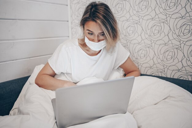 Mujer enferma en mascarilla protectora trabajando en una computadora portátil en la cama