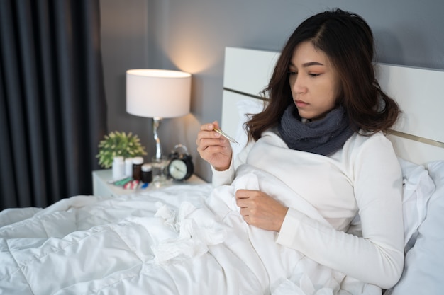 Mujer enferma que usa un termómetro para controlar su temperatura y su dolor de cabeza en la cama