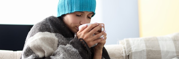 Mujer enferma con sombrero debajo de la manta sostiene una taza de medicina