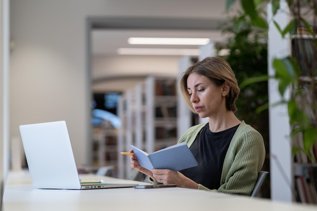 Mujer enfocada escribiendo una lista de tareas pendientes en un cuaderno mientras trabaja de forma remota en una computadora portátil en una biblioteca acogedora y tranquila