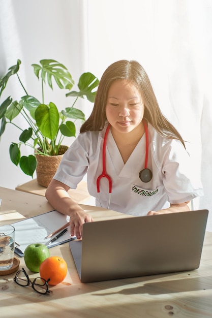 Mujer enfocada en uniforme médico sentada en una mesa de madera y mirando la pantalla de la computadora portátil mientras trabaja en una clínica moderna
