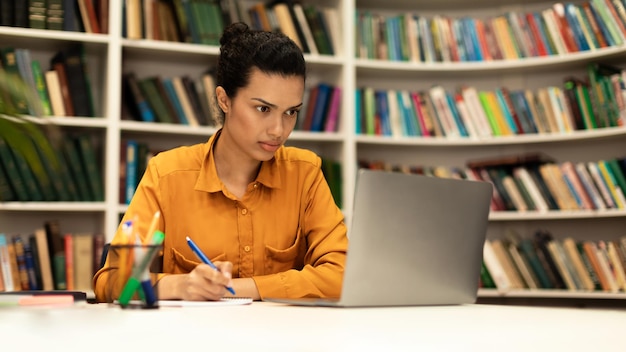 Mujer enfocada usando una computadora portátil y escribiendo en un cuaderno estudiando en línea en el panorama interior de la biblioteca con