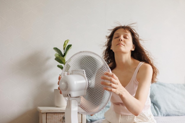 Foto mujer se enfría en casa con la ayuda de un ventilador de aire acondicionado se sienta en la cama frente a un eléctrico