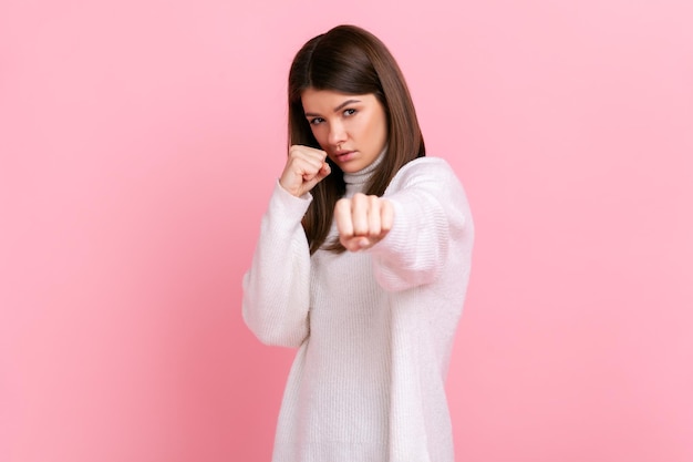 Mujer enojada agresiva con cabello oscuro lista para pelear, boxeando a la cámara con puños apretados, usando suéter blanco de estilo casual. Disparo de estudio interior aislado sobre fondo rosa.