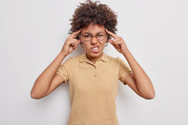 Mujer enojada disgustada aprieta los dientes y mantiene las manos en las sienes sufre de dolor de cabeza o migraña usa gafas transparentes redondas camiseta beige informal intenta concentrar las muecas del dolor