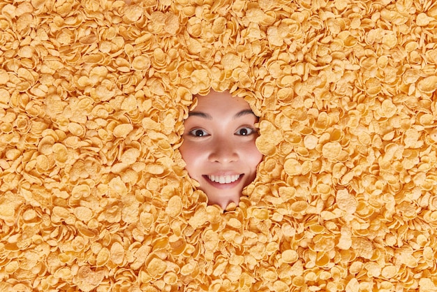 mujer enterrada en copos de maíz sonríe con los dientes con dientes blancos incluso está de buen humor se prepara para la hora del desayuno rodeada de cereales hace una foto creativa de la comida