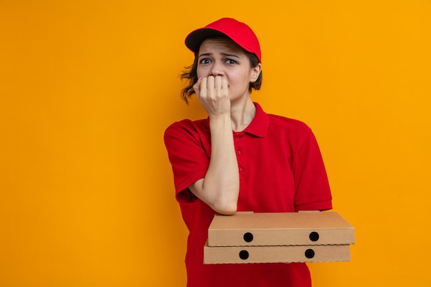 Mujer de entrega bonita joven ansiosa sosteniendo cajas de pizza y mordiéndose las uñas