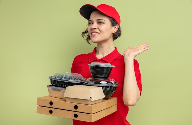 Mujer de entrega bonita joven complacida de pie con la mano levantada y sosteniendo contenedores de alimentos con envases en cajas de pizza