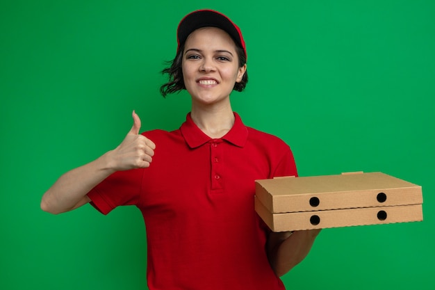 Mujer de entrega bonita joven sonriente sosteniendo cajas de pizza y pulgar hacia arriba