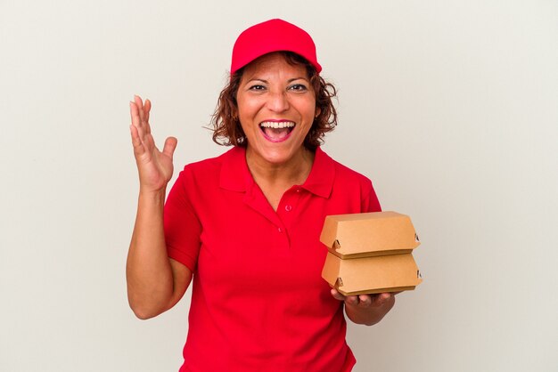 Mujer de entrega de mediana edad tomando burguers aislado sobre fondo blanco recibiendo una agradable sorpresa, emocionado y levantando las manos.