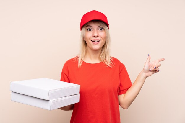 Mujer de entrega de pizza sosteniendo una pizza sobre pared aislada sorprendido y apuntando con el dedo al lado