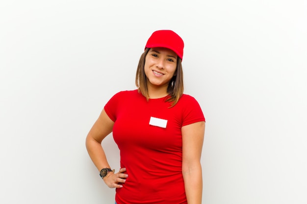 Mujer de entrega sonriendo felizmente con una mano en la cadera y actitud segura, positiva, orgullosa y amigable contra la pared blanca