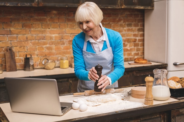 La mujer envejecida mayor atractiva está cocinando en cocina. Abuela haciendo sabrosa hornada. Usando la computadora portátil.