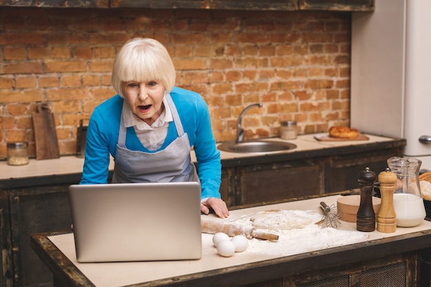 La mujer envejecida mayor sorprendida sorprendida atractiva está cocinando en cocina. Abuela haciendo sabrosa hornada. Usando la computadora portátil.