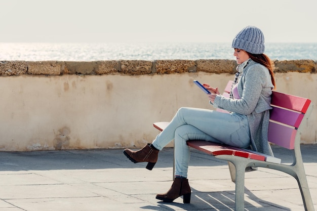 Mujer envía un mensaje por teléfono móvil sentada en un banco de colores con el mar de fondo Estilo de vida