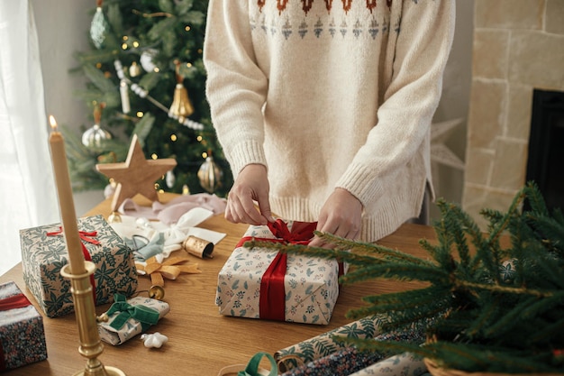 Mujer envolviendo un elegante regalo de Navidad con cinta roja en una mesa de madera con decoraciones festivas en una habitación escandinava decorada Feliz Navidad y Felices Fiestas