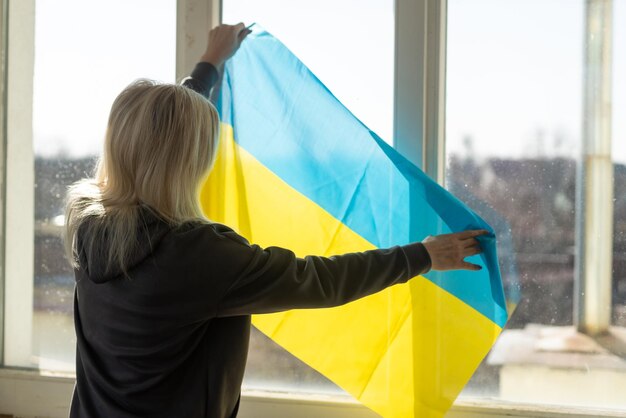 Mujer envuelta en la bandera de Ucrania mirando por la ventana. copie el espacio