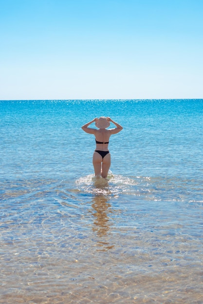 Una mujer esbelta madura en traje de baño entra en el mar, vista trasera. Vacaciones de verano, viajes y turismo.