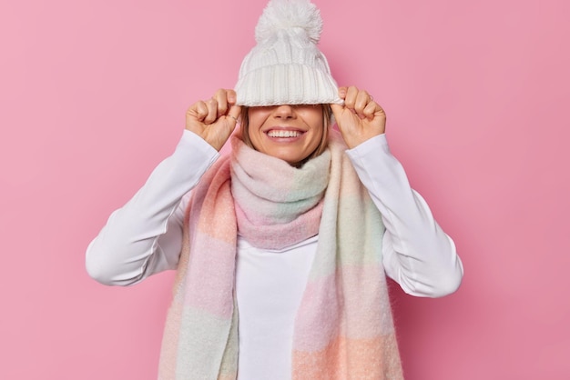 La mujer esconde los ojos con un sombrero blanco de punto viste un jersey y una bufanda cálida alrededor del cuello vestidos para el clima invernal se siente feliz aislado en rosa. Concepto de temporada y ropa.