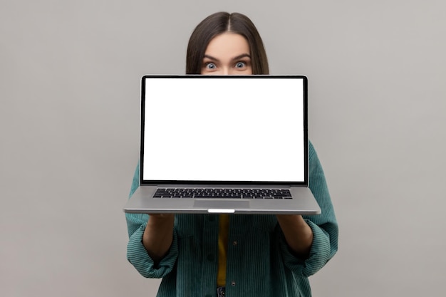 Mujer escondiendo la mitad de la cara detrás de la computadora portátil con la pantalla vacía mirando a la cámara con grandes ojos sorprendidos