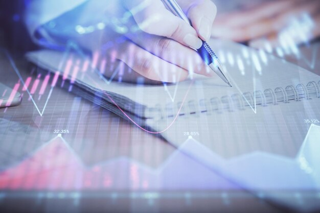 Una mujer escribe información sobre el mercado de valores en el bloc de notas. Hologramas de gráficos Forex delante Concepto de investigación Exposición múltiple