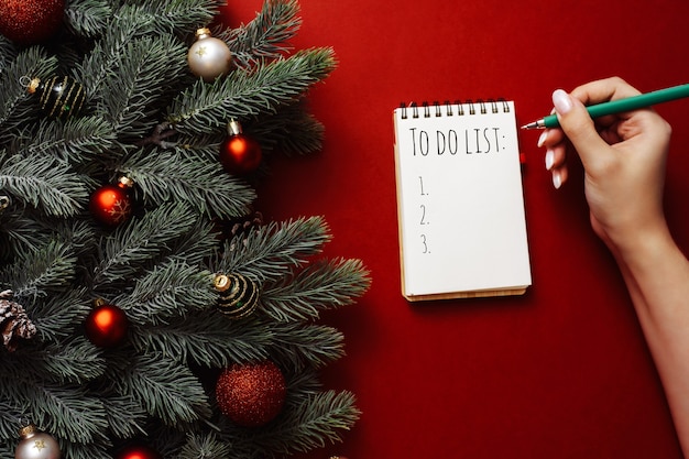 Una mujer escribe una lista de compras o de hacer en un cuaderno sobre un fondo rojo junto a las ramas y adornos de los árboles de Navidad.