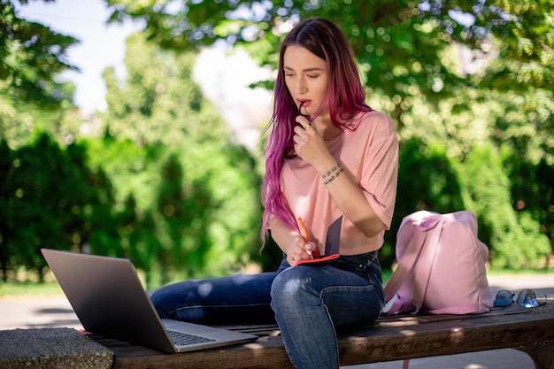 Mujer escribiendo en un cuaderno sentado en un banco de madera en el parque. Chica trabajando al aire libre en una computadora portátil, copie el espacio.