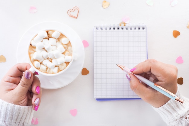 Mujer escribiendo en un cuaderno y sosteniendo una taza de chocolate caliente con malvaviscos