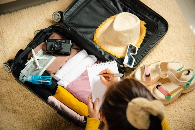 Foto mujer escribiendo papel tome nota y empacando maleta para vacaciones escribiendo lista de papel sentado en la habitación