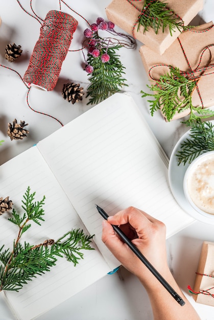 Mujer escribir deseos o hacer una lista en el cuaderno, taza de café, regalo de Navidad o caja de regalo, decorada con ramas de árboles de navidad, piñas, bayas rojas, en la mesa de mármol blanco, vista superior del espacio de copia