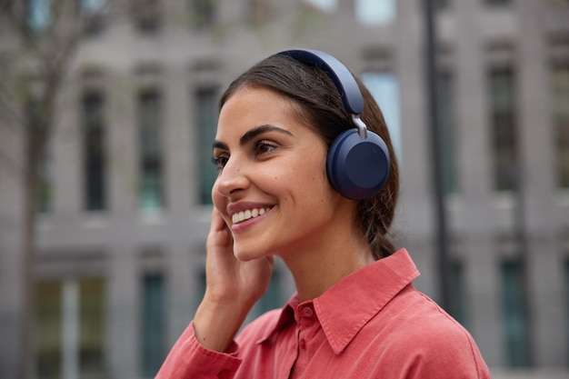 Mujer escucha música en auriculares al aire libre sonríe suavemente enfocado hacia adelante disfruta de su canción favorita viste camisa roja posa contra borrosa