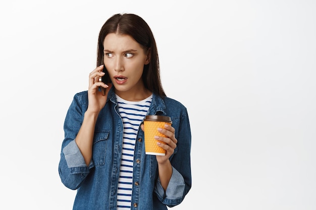 Una mujer escucha noticias impactantes a través de una llamada telefónica, sostiene una taza de café amarilla para llevar del café, habla por teléfono con cara preocupada, está ocupada contra el fondo blanco