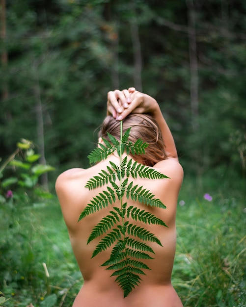 La mujer con la espalda desnuda se sienta en el bosque y sostiene una hoja de helecho verde en el bosque