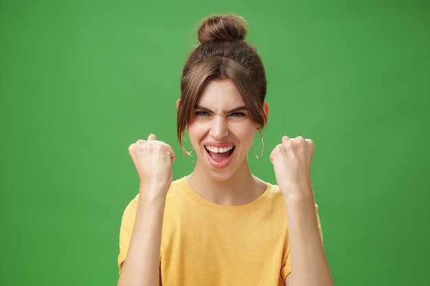 Mujer con espíritu de ganador levantando los puños cerrados sonriendo entusiasmados y animando de apoyo estando lista para el gimnasio ejercitó boosing confidentes con gritos sobre la pared verde.