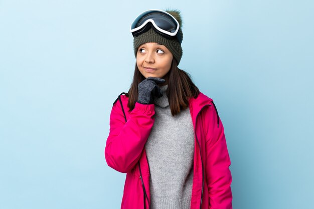 Mujer de esquiador de raza mixta con gafas de snowboard sobre espacio azul aislado pensando una idea mientras mira hacia arriba