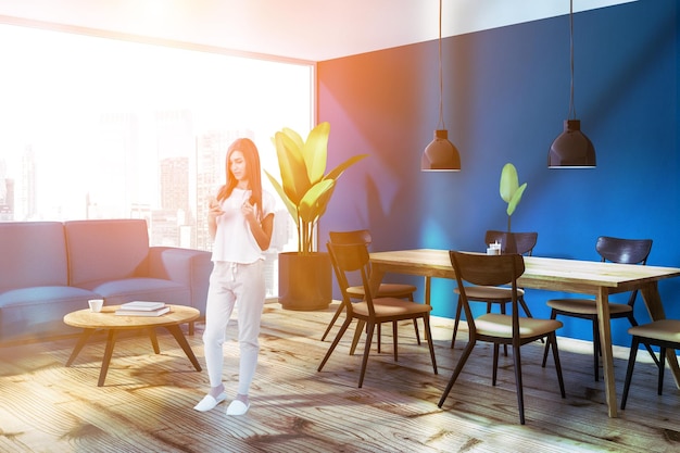 Mujer en la esquina del comedor loft con paredes azules, suelo de madera, mesa de madera con sillas azules y de madera y sofá azul cerca de la mesa baja. imagen tonificada