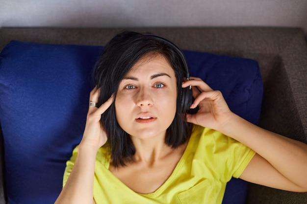 La mujer está acostada en el sofá y escuchando música. Mujer alegre en ropa casual con auriculares y teléfono inteligente, escuchando podcast en el sofá en casa.