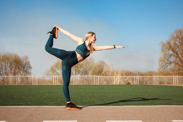 La mujer está estirando la espalda y la pierna, de pie en la pose del señor de la danza al aire libre en el estadio