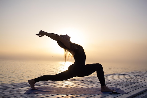 una mujer está haciendo yoga en un muelle con el sol detrás de ella
