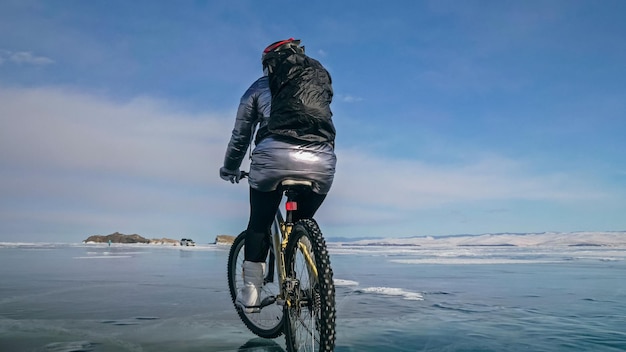La mujer está montando en bicicleta en el hielo. La niña está vestida con una chaqueta plateada, una mochila y un casco. Hielo del lago Baikal congelado. Los neumáticos en bicicleta están cubiertos de púas.