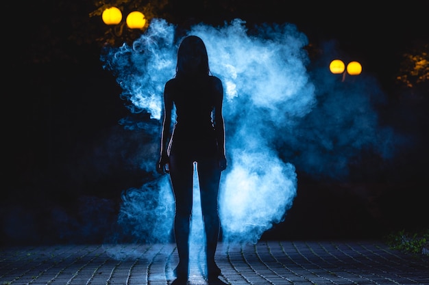 La mujer está parada en la calle oscura sobre el fondo de humo azul. Noche