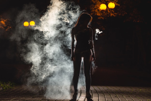 La mujer está parada en el callejón oscuro sobre el fondo de humo. Noche