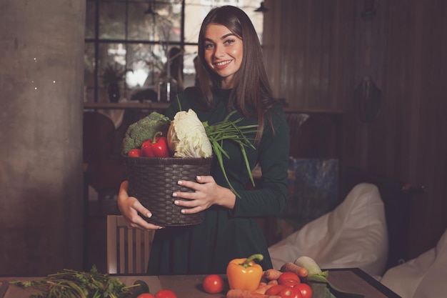 La mujer está de pie con una cesta de verduras cerca de la mesa de madera.
