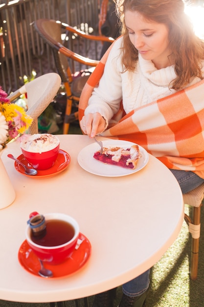 La mujer está sentada en la cafetería al aire libre y come pastel y cacao, otoño acogedor