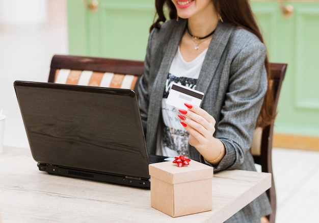 La mujer está sentada en la cafetería, sosteniendo la tarjeta de crédito y la caja roja actual, fácil de ordenar, comprar en línea, mirando a la cámara. Interior. Foto horizontal