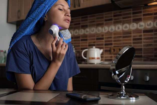 Una mujer está sentada en la cocina y está haciendo un procedimiento de belleza usando un aparato de belleza.