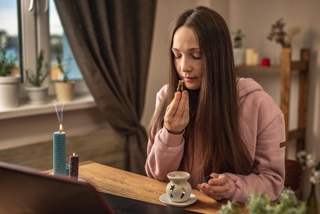 Una mujer está sentada frente a una computadora portátil escuchando a un instructor o una videoconferencia sobre meditación y olfateando aceite esencial de una botella Concepto de terapia a distancia y sesión de meditación