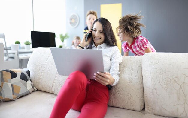 La mujer está sentada en el sofá sosteniendo una computadora portátil y hablando por teléfono inteligente al lado de los niños están jugando