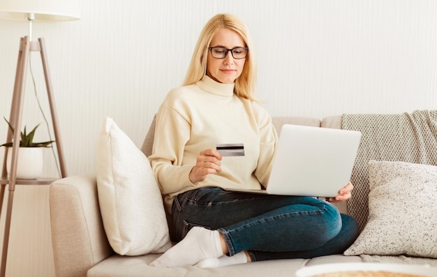 Una mujer está sentada en un sofá usando una computadora portátil y sosteniendo una tarjeta de crédito que parece estar comprando