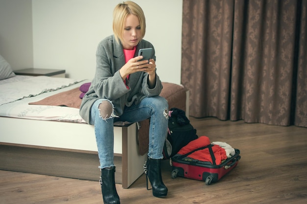 Una mujer está sentada con un teléfono en la cama cerca de la maleta.